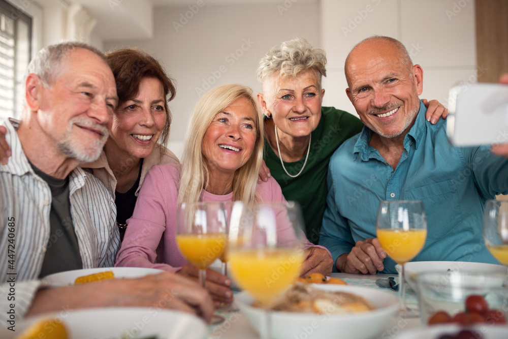 Group of senior friends having party indoors, taking selfie when eating at the table.