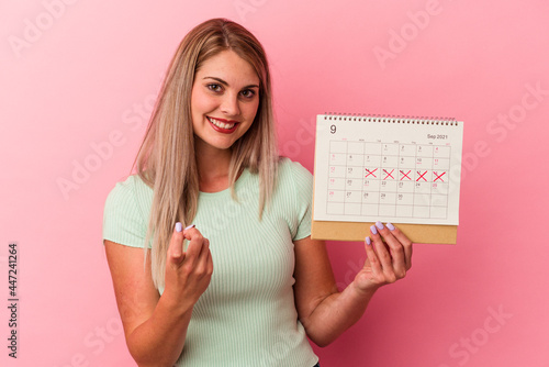 Young caucasian woman holding a calendar isolated on pink background pointing with finger at you as if inviting come closer.