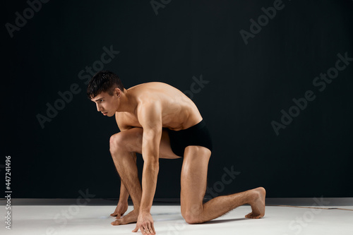 handsome man muscularly muscled in black panties stands on his knee dark background