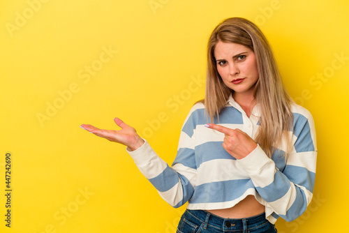 Young russian woman isolated on yellow background excited holding a copy space on palm. © Asier