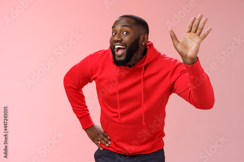 Attractive funny african american gay beard in red hoodie raise palm waving hello hi howdy gesture acting feminine cute fool around welcoming guests greeting standing pink background photo