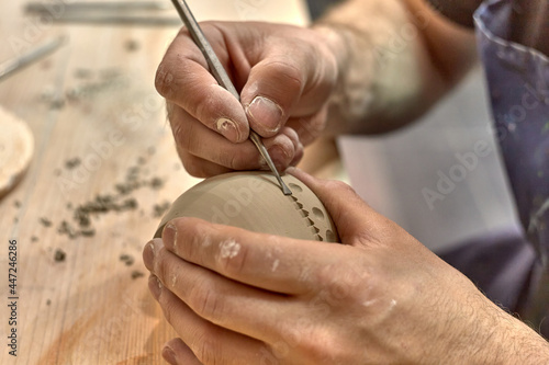 Hands of a ceramist who carves a round ornament on a cup made on a potter's wheel with a knife