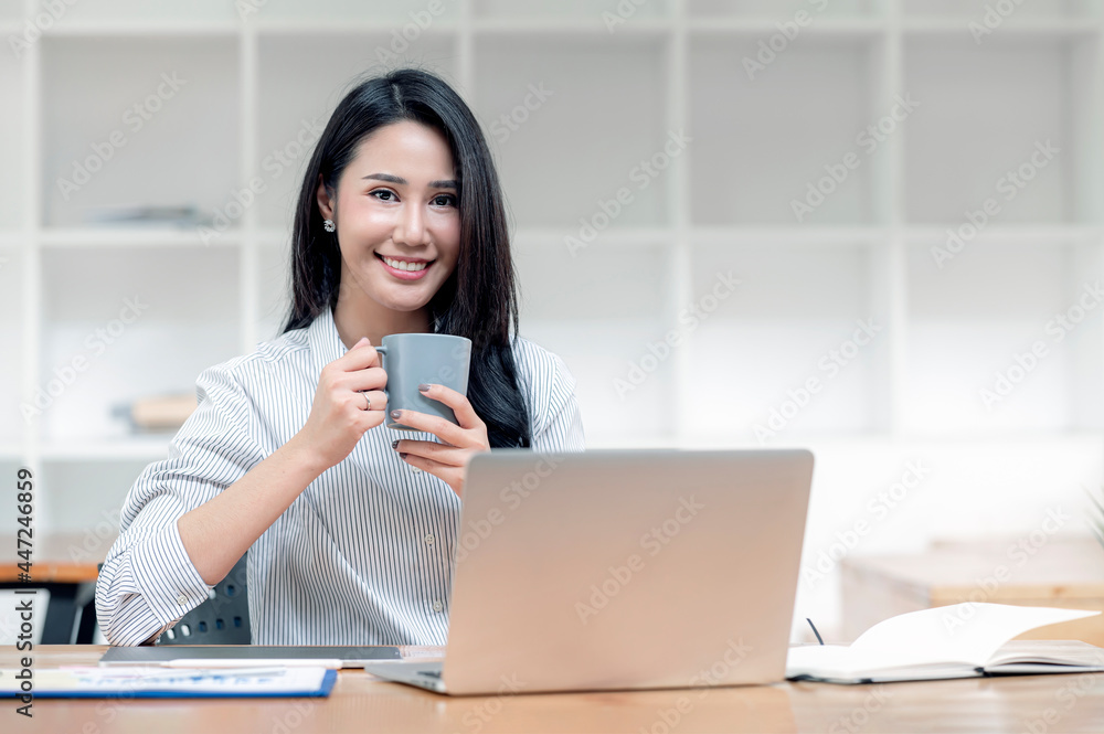 Portrait of young asian woman holding mug, smiling and looking at camera while sitting at the table with laptop in living room. Portrait of young asian woman holding mug, smiling and looking at camera while sitting at the table with laptop in living room.