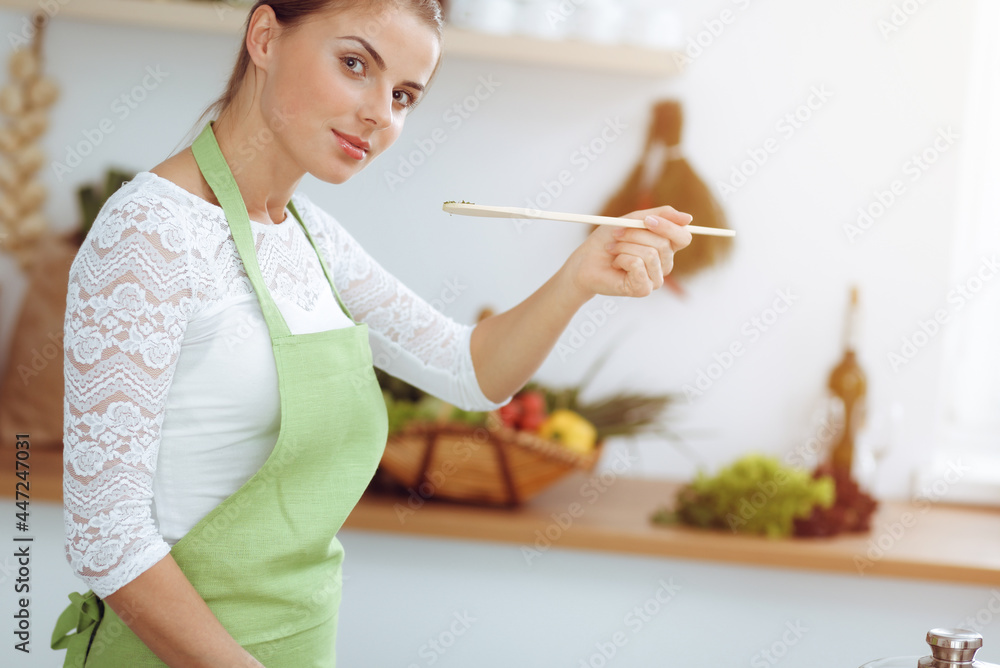 An attractive young dark-haired woman frying meal at worm pan by new keto recipe while standing and smiling in sunny kitchen. Cooking and householding concepts An attractive young dark-haired woman frying meal at worm pan by new keto recipe while standing and smiling in sunny kitchen. Cooking and householding concepts