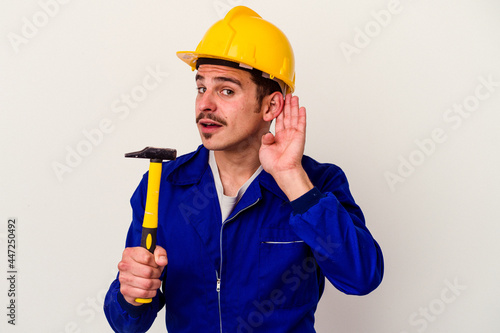 Young caucasian worker man holding a hammer isolated on white background trying to listening a gossip.