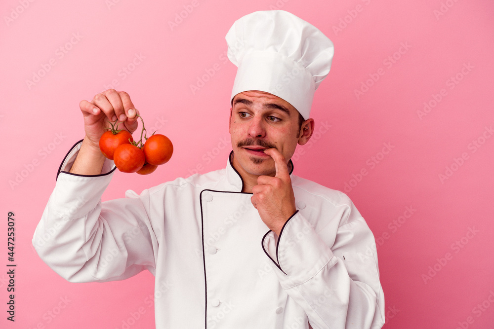 Young caucasian chef man holding tomatoes isolated on pink background relaxed thinking about something looking at a copy space. Young caucasian chef man holding tomatoes isolated on pink background relaxed thinking about something looking at a copy space.