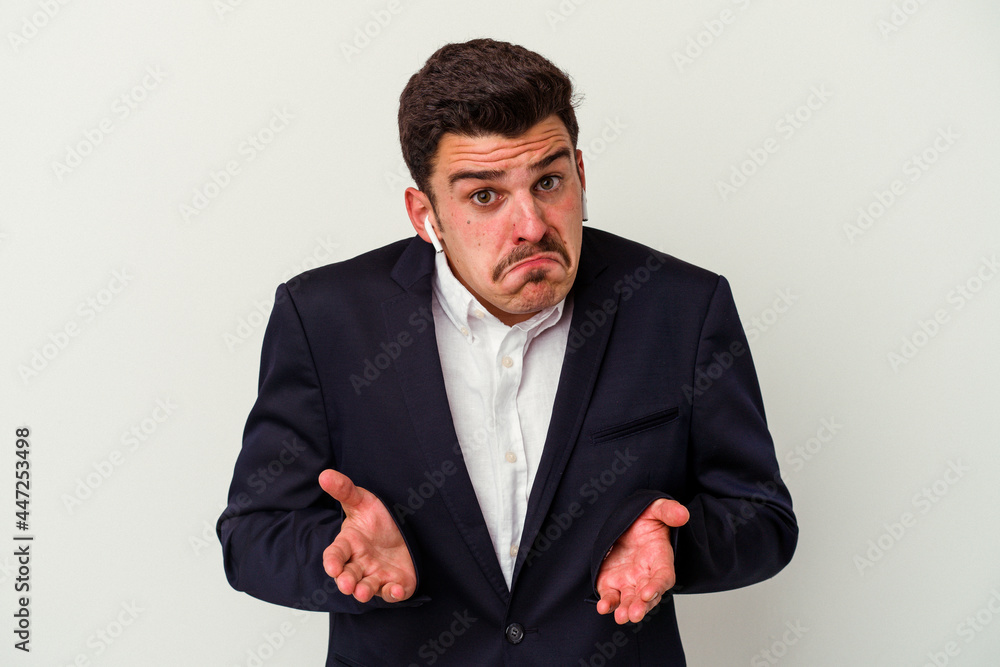 Young business caucasian man wearing wireless headphones isolated on white background shrugs shoulders and open eyes confused. Young business caucasian man wearing wireless headphones isolated on white background shrugs shoulders and open eyes confused.