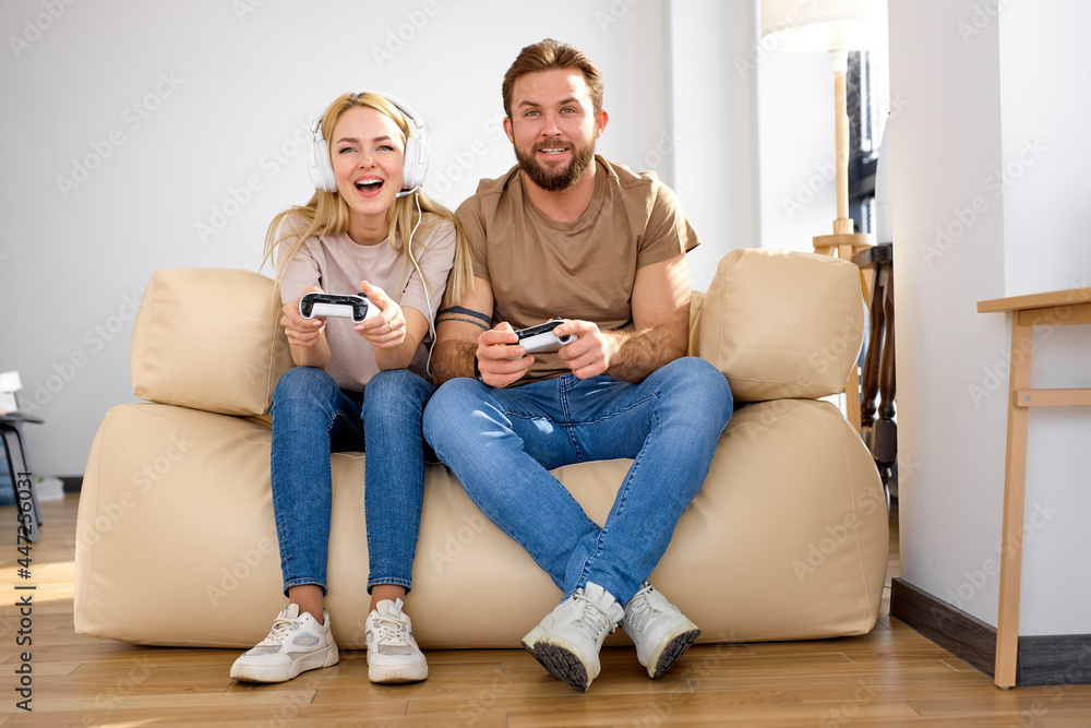 Overjoyed caucasian couple playing video game, holding remote controllers, sitting on cozy couch at home, happy laughing man and woman enjoying leisure time with console, relaxing, having fun together