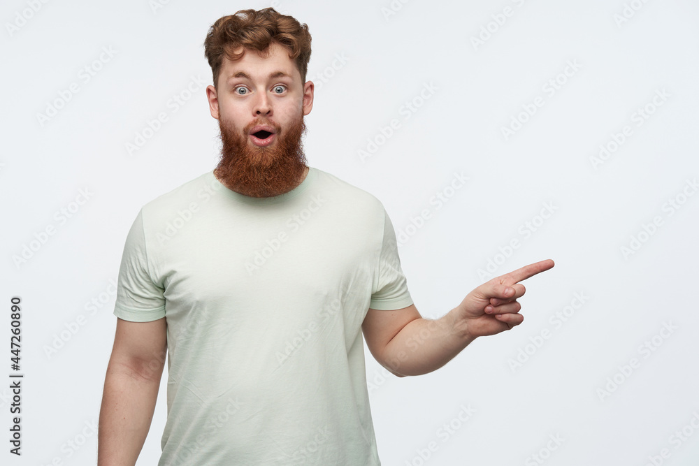 indoor shot of young bearded redhead guy, wears blank t-shirt, pointing with a finger at right side copy space, smiling and has amazed facial expression. isolated over white background. indoor shot of young bearded redhead guy, wears blank t-shirt, pointing with a finger at right side copy space, smiling and has amazed facial expression. isolated over white background.