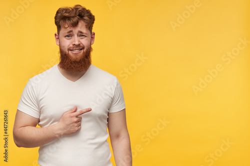 confused guy with red hair and beard wears blank t-shirt, watching into camera and pointing to he right side at copy space with a disgust facial expression , isolated over yellow background