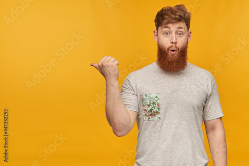 indoor shot of amazed, positive young bearded male with red hair, pointing with a finger left at copy space with amazed facial expression. Isolated over yellow background