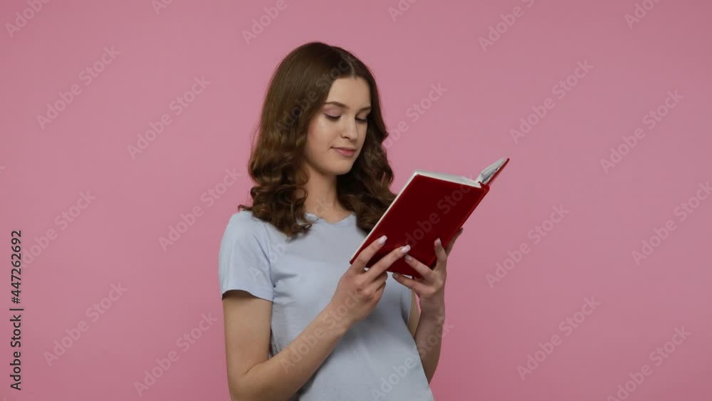 Adorable smart young female student in blue casual T-shirt, reading book showing thumbs up, recommend book with satisfied with plot. Indoor studio shot isolated over pink background. Adorable smart young female student in blue casual T-shirt, reading book showing thumbs up, recommend book with satisfied with plot. Indoor studio shot isolated over pink background.