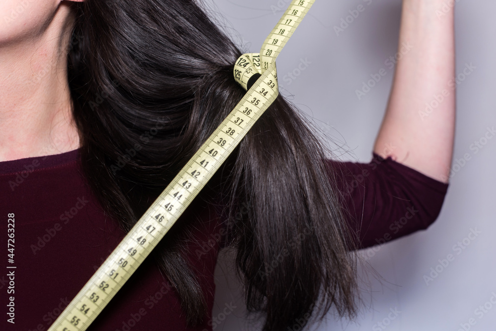 Detalle de una mujer sosteniendo una cinta métrica amarilla alrededor de un mechón de su cabello largo. Primer plano de una cinta de medir con números que se muestran. - obrazy, fototapety, plakaty 