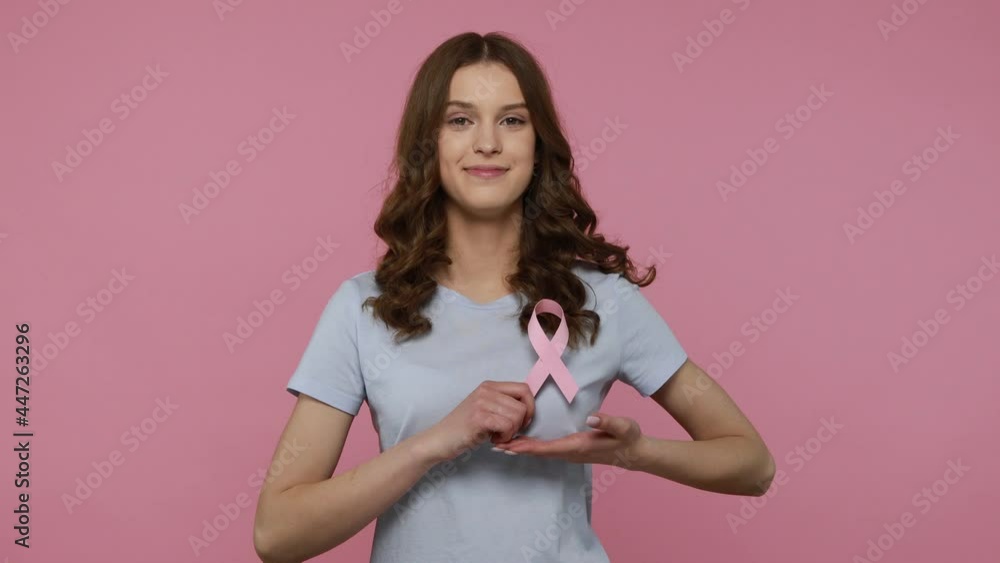 Adorable woman with wavy hair in casual T-shirt showing pink ribbon, symbol of breast cancer awareness, female health diagnostics, disease prevention. Indoor studio shot isolated over pink background. Adorable woman with wavy hair in casual T-shirt showing pink ribbon, symbol of breast cancer awareness, female health diagnostics, disease prevention. Indoor studio shot isolated over pink background.