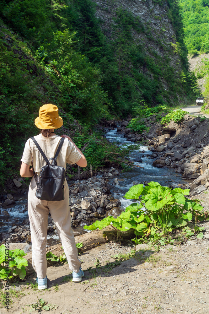 Traveler in a hat and a backpack is resting in nature. A tourist girl looks at the panorama of a mountain river. Mountain beautiful landscape Traveler in a hat and a backpack is resting in nature. A tourist girl looks at the panorama of a mountain river. Mountain beautiful landscape
