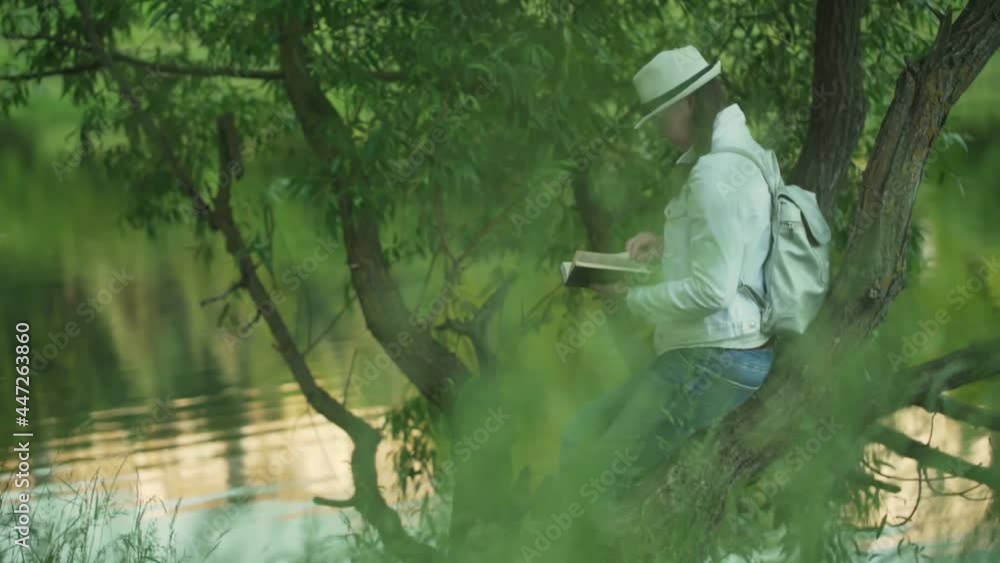 Lady brunette sits on green tree and reads fiction book by calm lake with landscape reflections on sunny summer day slow motion