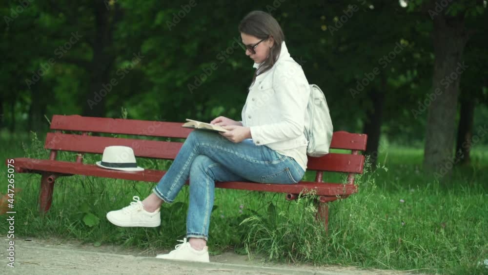 Young woman brunette sits on red wooden bench in park near hat enjoys nature with green trees holding small book slow motion Young woman brunette sits on red wooden bench in park near hat enjoys nature with green trees holding small book slow motion