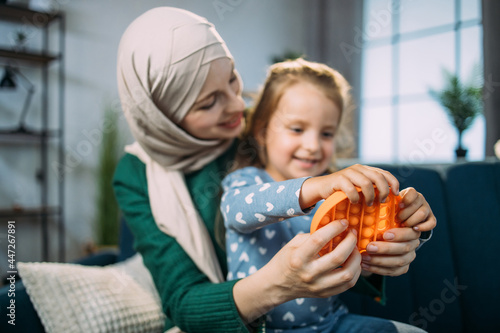 Orange antistress sensory toy fidget push pop it. Beautiful Muslim woman in hijab  sitting on sofa at home and playing with colorful pop it fidget toy with her little cute daughter. Focus on poppit