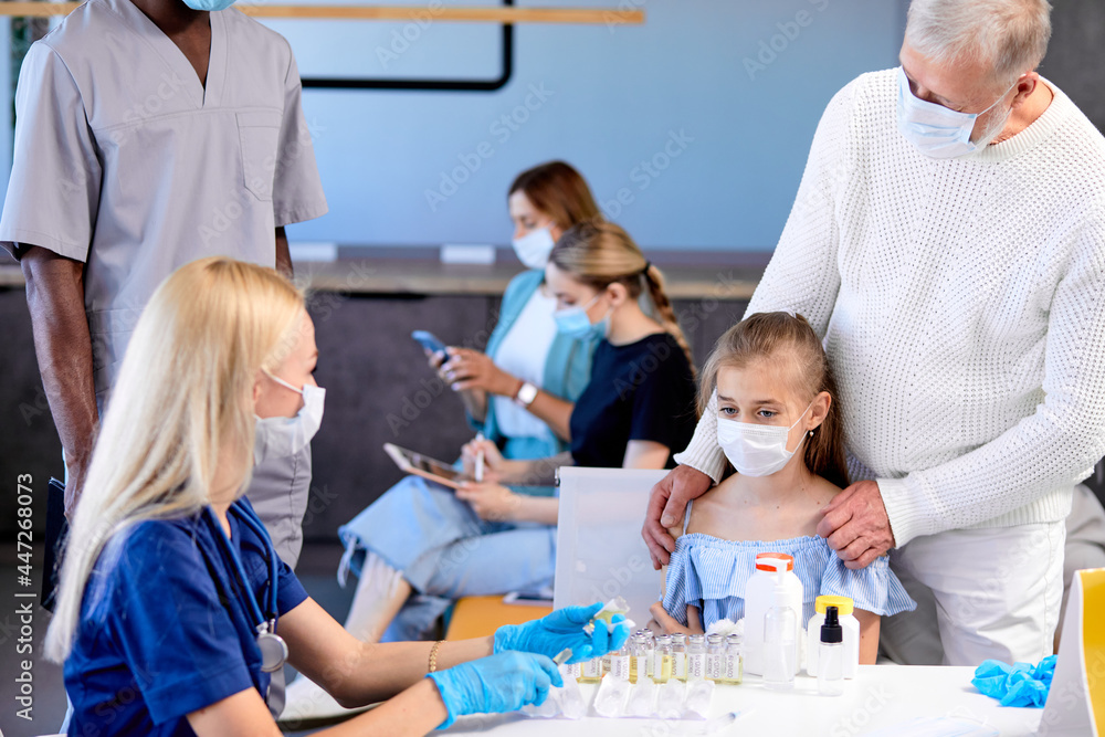 Mass immunization, scared child girl will get vaccination in hospital. Professional adult female nurse or doctor in gloves dials vaccine into syringe to inject to patient. focus on girl came with dad Mass immunization, scared child girl will get vaccination in hospital. Professional adult female nurse or doctor in gloves dials vaccine into syringe to inject to patient. focus on girl came with dad