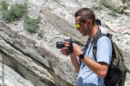 A young male tourist in sports glasses stands with a camera in his hands against a background of mountains and greenery