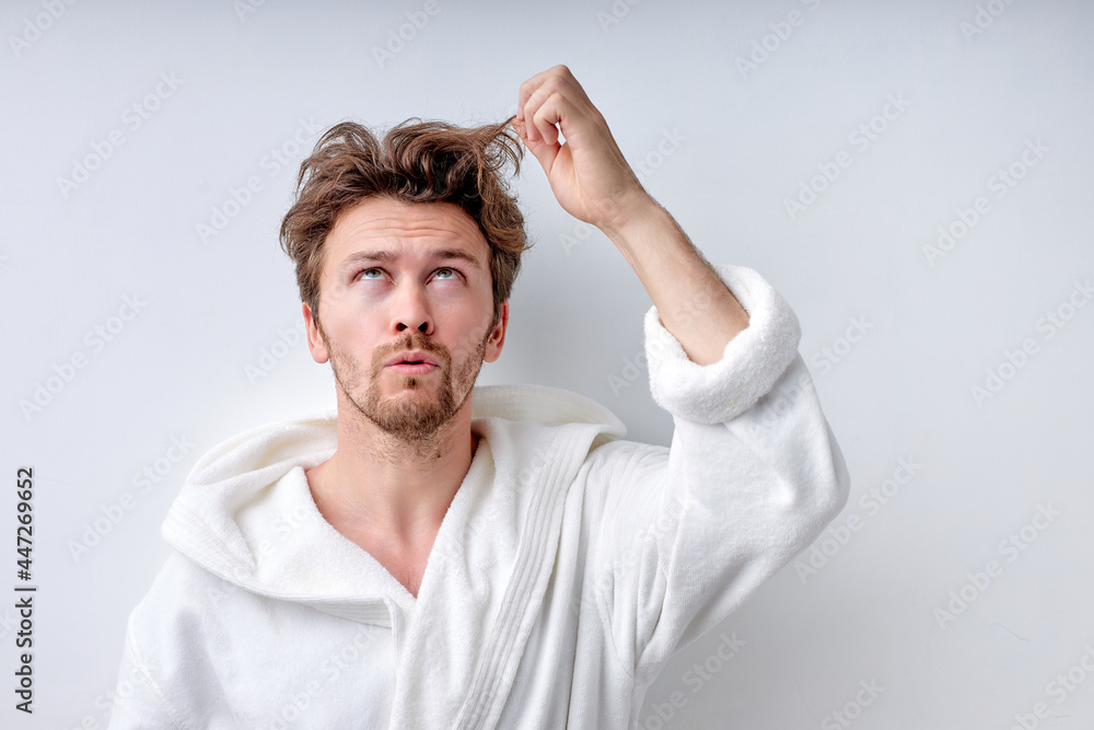 shaggy young caucasian guy in white bathrobe isolated on white background, touches long regrown hair and looks up with sad face, thinking. Beauty, people lifestyle concept. Copy space. Hairstyle shaggy young caucasian guy in white bathrobe isolated on white background, touches long regrown hair and looks up with sad face, thinking. Beauty, people lifestyle concept. Copy space. Hairstyle