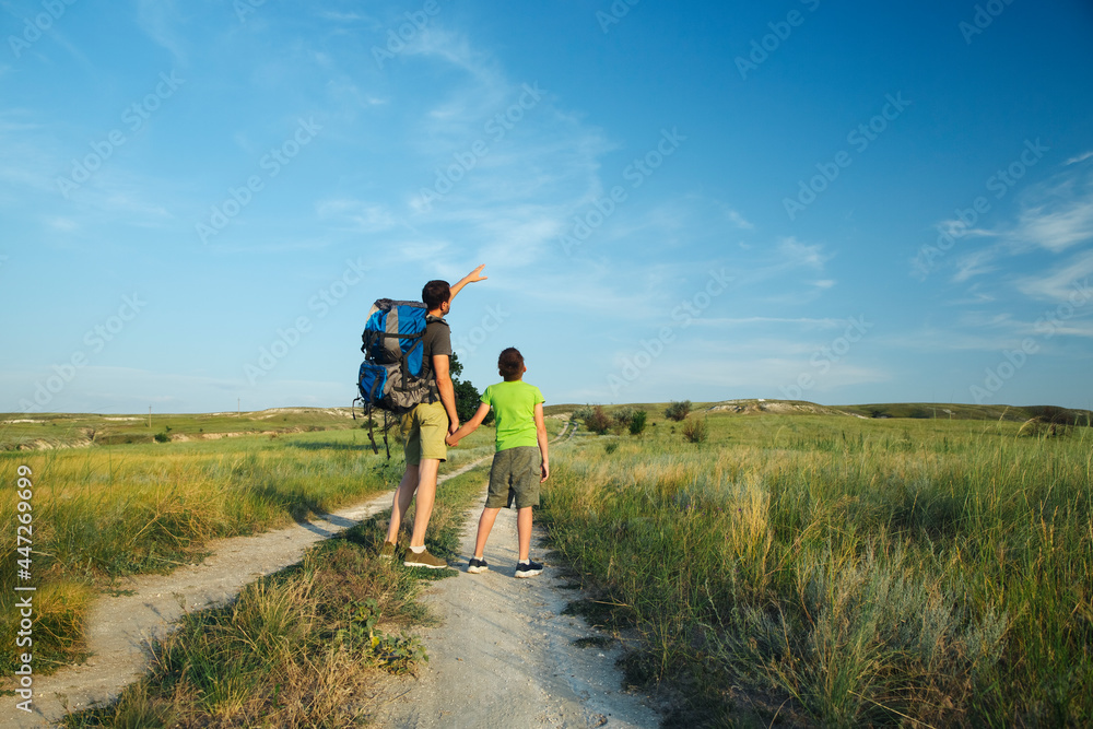 A young dad holds his little son by the hand and points to the sky into the distance, standing on the big road, traveling with a backpack through the highlands