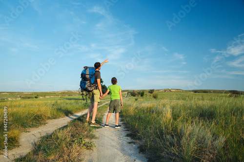A young dad holds his little son by the hand and points to the sky into the distance, standing on the big road, traveling with a backpack through the highlands