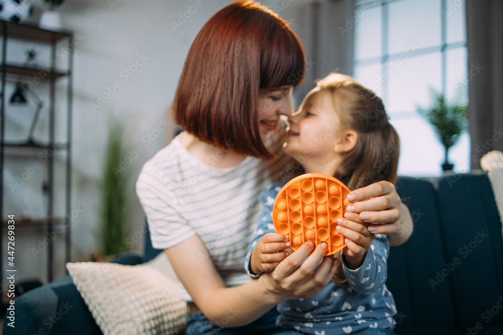 Trendy toy antistress, trendy pop it fidget toy. Likable family, pretty mom and cute little daughter, sitting on the sofa at home and touching noses, playing pop pit game Trendy toy antistress, trendy pop it fidget toy. Likable family, pretty mom and cute little daughter, sitting on the sofa at home and touching noses, playing pop pit game