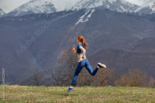 Side View Portrait Of Athlete Redhead Lady Jumping High During Training In Nature, Sportswoman In Blue Sportive Outfit Enjoy Jogging, Mountains Landscape In The Background