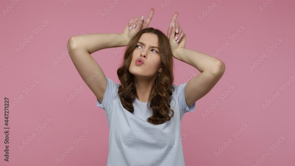 Playful teenager girl with wavy hair wearing blue T-shirt making bull horn gesture, pretending to be dangerous, woman showing defence herself. Indoor studio shot isolated over pink background. Playful teenager girl with wavy hair wearing blue T-shirt making bull horn gesture, pretending to be dangerous, woman showing defence herself. Indoor studio shot isolated over pink background.