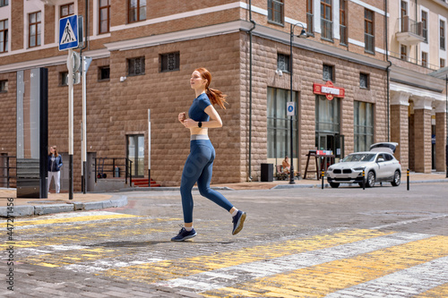 Young woman running on road in city. Lifestyle morning run. Fitness and healthy lifestyle concept. Side view portrait on redhead lady in blue sportswear engaged in sport, jogging outdoors © Roman