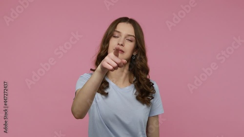 Beautiful young lady in T-shirt showing loser gesture and points finger to camera, expressing disrespect, accusing blaming for failure, life troubles. Indoor studio shot isolated over pink background. photo