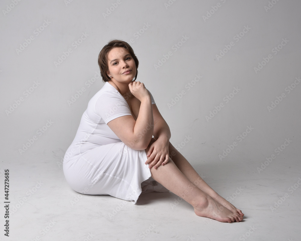 Obraz premium Full length portrait of young plus sized woman with short brunette hair, wearing l tight white body con dress, kneeling pose with gestural hands on ground with light studio background.