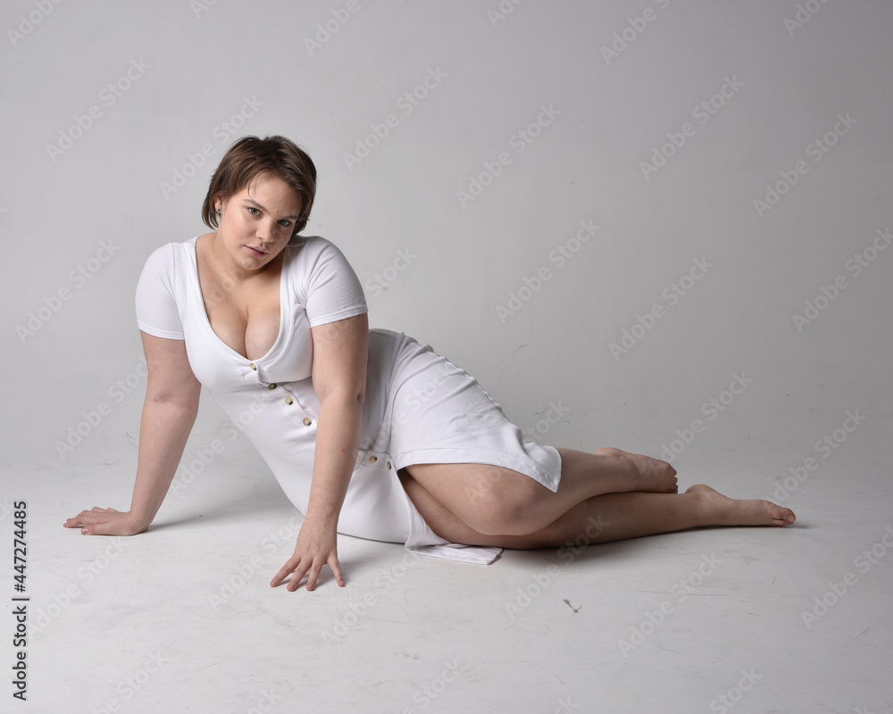 Full length portrait of young plus sized woman with short brunette hair, wearing l tight white body con dress, kneeling pose with gestural hands on ground with light studio background. Full length portrait of young plus sized woman with short brunette hair, wearing l tight white body con dress, kneeling pose with gestural hands on ground with light studio background.