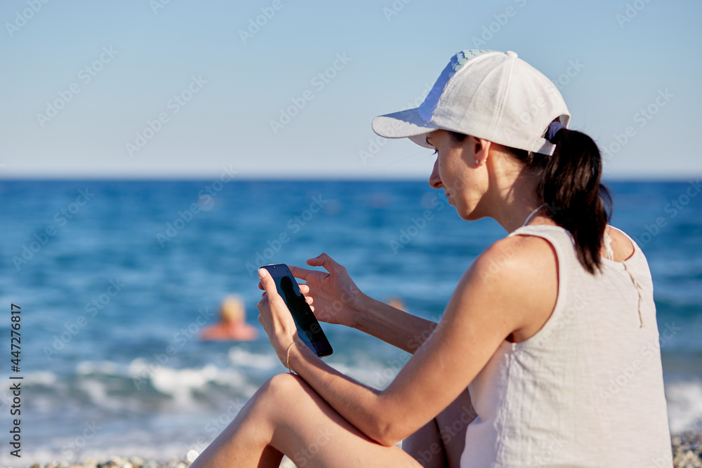  Young woman is sitting in a hat with a phone on the beach near the sea is making a selfie. Resting, traveling and relaxing concept. Copy space