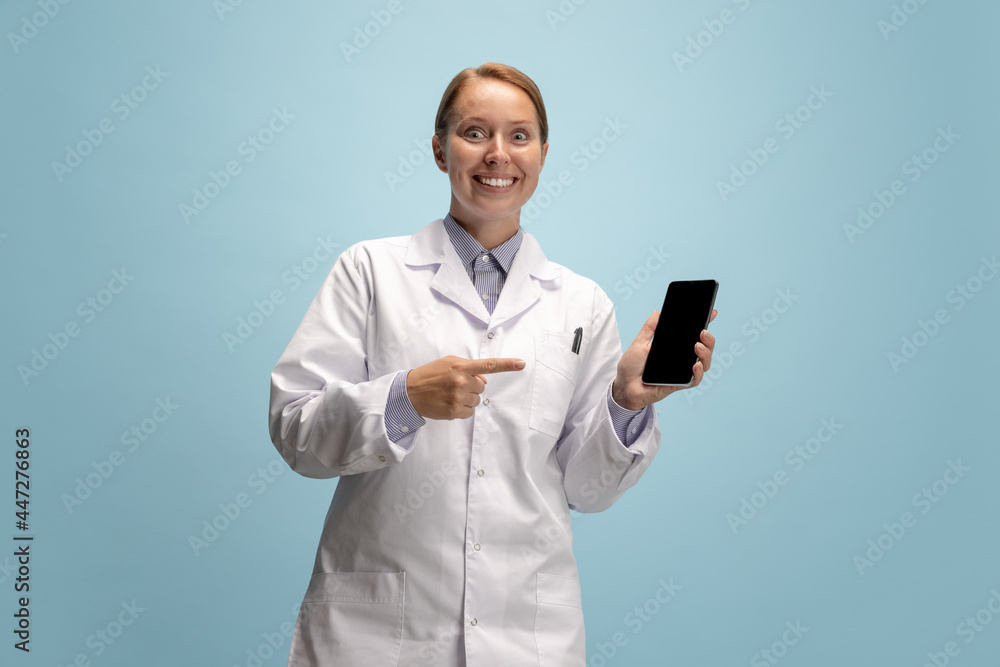 Portrait of female doctor, therapeutic or medical advisor with smartphone screen isolated on blue background. Funny happy emotions. Concept of healthcare, care medicine and humor Portrait of female doctor, therapeutic or medical advisor with smartphone screen isolated on blue background. Funny happy emotions. Concept of healthcare, care medicine and humor
