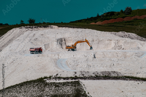 Heavy machinery an excavator and a KAMAZ truck carry out mining on a white limestone quarry under the blue sky on a working day