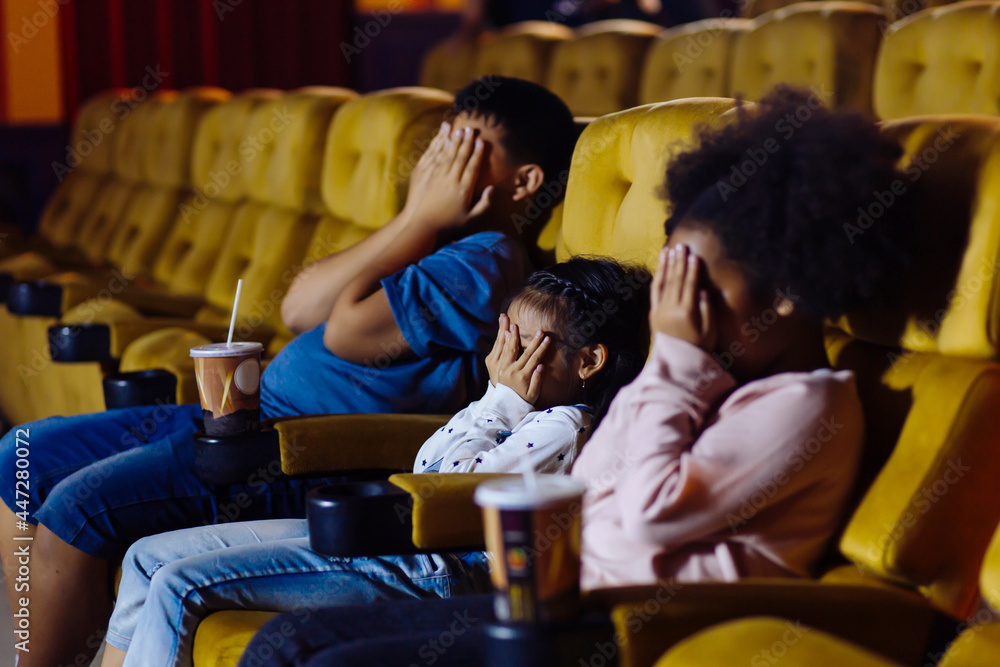 Young boy and girls sitting in movie theater with cold drinks closing their eyes with hands
