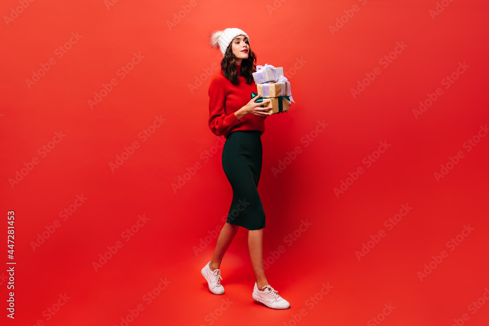 Full-length portrait of girl holding gift box. Charming woman in black skirt and bright sweater walks on red background. Full-length portrait of girl holding gift box. Charming woman in black skirt and bright sweater walks on red background.