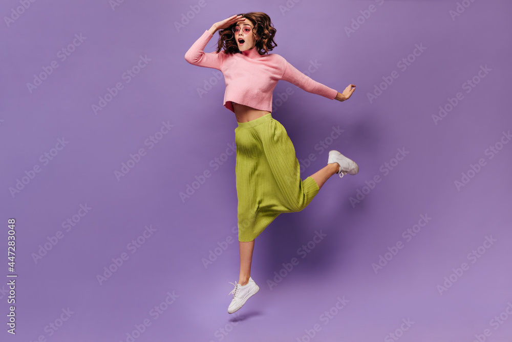 Curly woman jumping on purple background. Young cheerful girl in green skirt and pink sweater looks away on isolated. Curly woman jumping on purple background. Young cheerful girl in green skirt and pink sweater looks away on isolated.