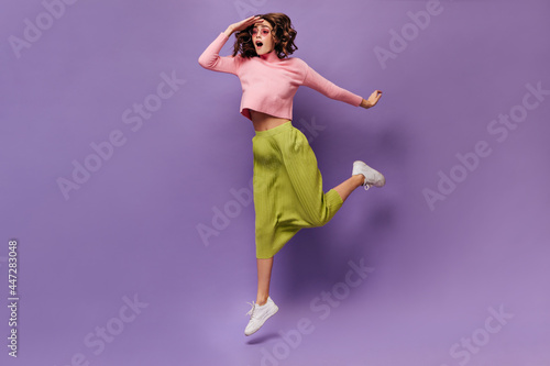 Curly woman jumping on purple background. Young cheerful girl in green skirt and pink sweater looks away on isolated.