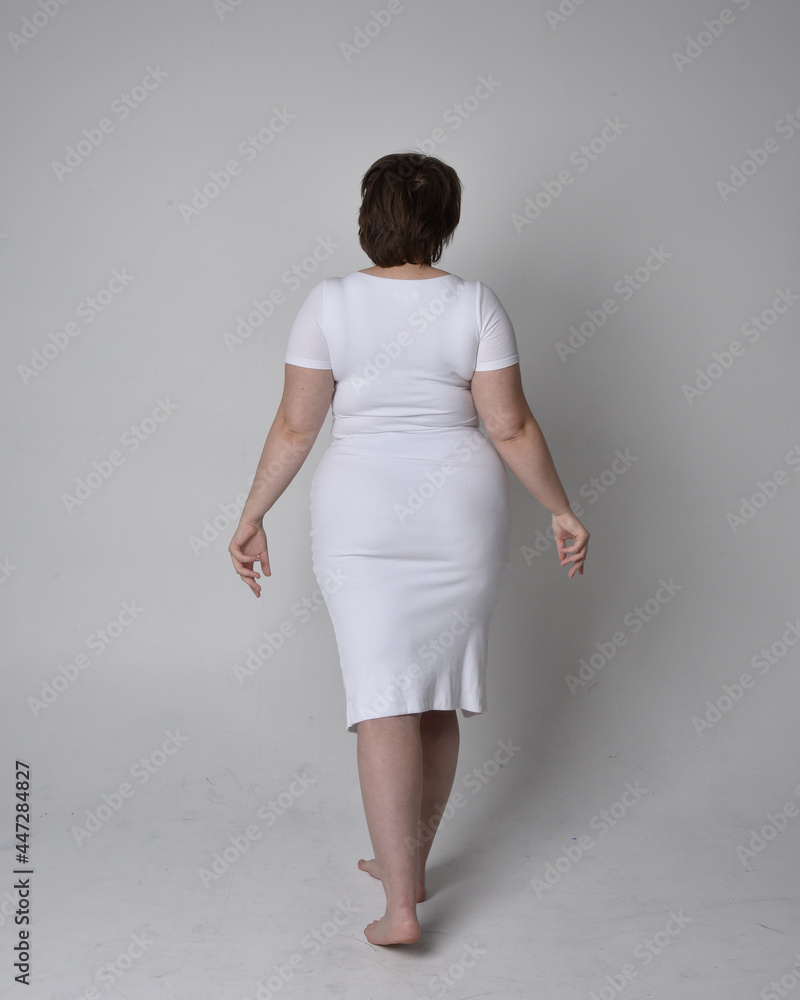 Full length portrait of young plus sized woman with short brunette hair, wearing a tight white body con dress, standing walking posewith light studio background. Full length portrait of young plus sized woman with short brunette hair, wearing a tight white body con dress, standing walking posewith light studio background.
