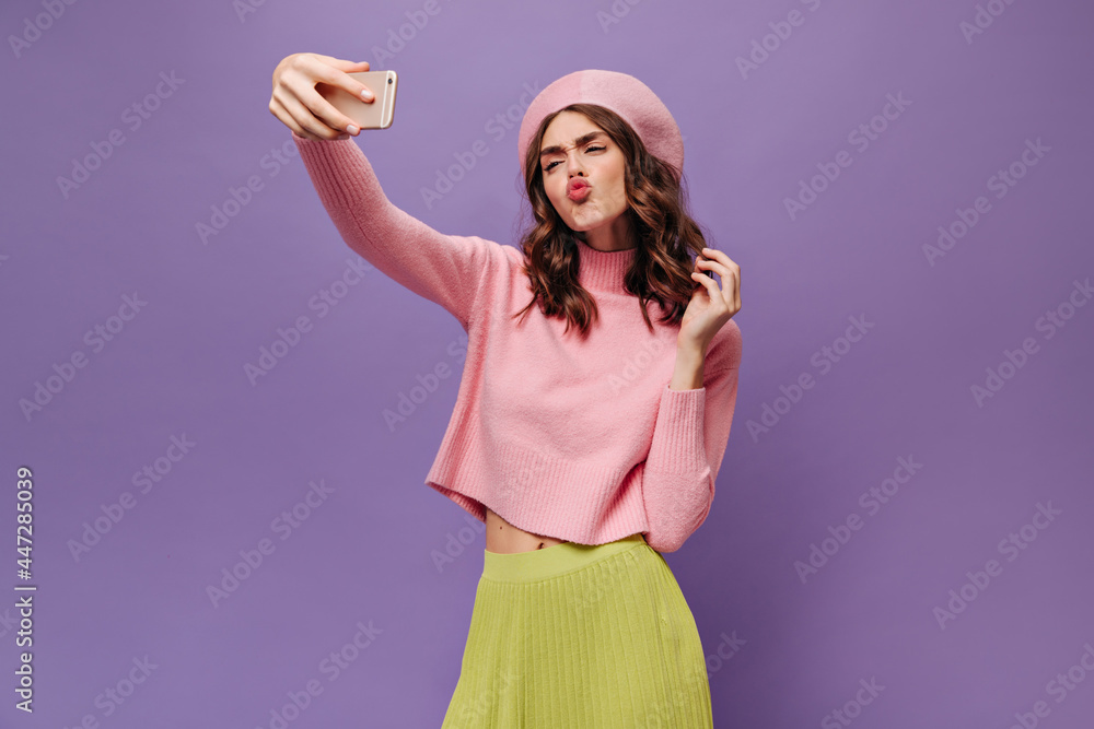 Attractive curly woman in green skirt, pink beret and pink sweater blows kiss, takes selfie and holds phone on purple background. Attractive curly woman in green skirt, pink beret and pink sweater blows kiss, takes selfie and holds phone on purple background.