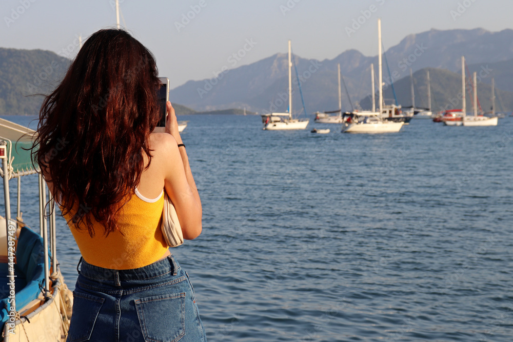 Beach vacation on the sea, summer leisure and travel. Girl in jeans photographing on smartphone camera yachts and mountains in mist Beach vacation on the sea, summer leisure and travel. Girl in jeans photographing on smartphone camera yachts and mountains in mist