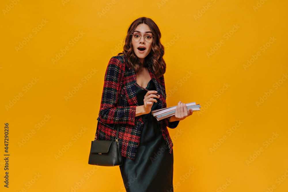 Surprised woman holds books. Beautiful curly lady in checkered jacket and dress looks into camera on orange background. Surprised woman holds books. Beautiful curly lady in checkered jacket and dress looks into camera on orange background.