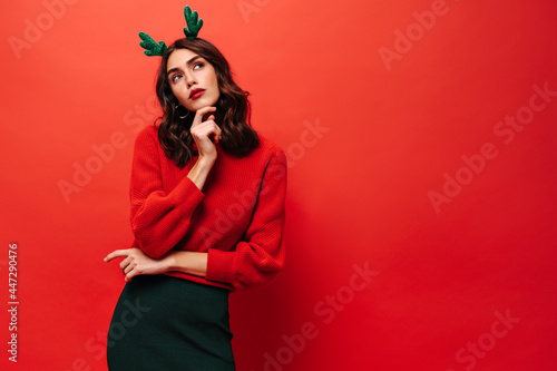 Pensive lady with brunette hair in red sweater and green skirt looking away on isolated backdrop. Woman in cool outfit posing on bright background..