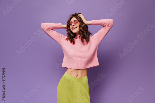 Curly girl ruffles hair on isolated. Cool charming woman in pink sweater, green skirt and sunglasses smiles on purple background.