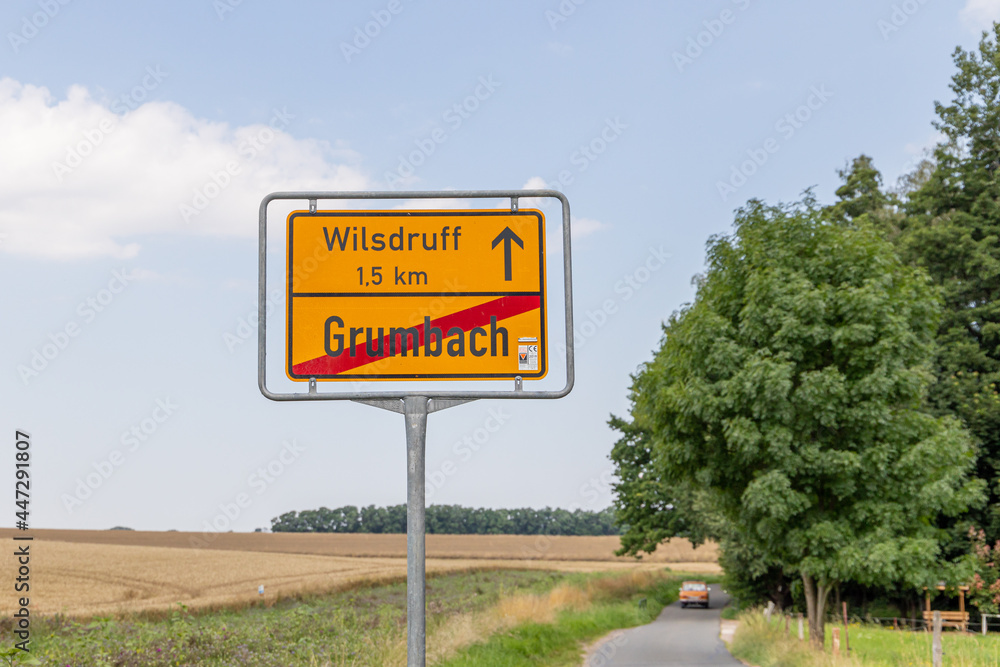 Yellow street sign or place name sign with Wilsdruff and Grumbach in German language. Grumbach is crossed out and in the background you can see a car driving. 