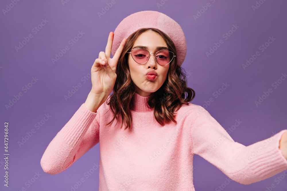 Naklejka premium Excited young woman takes selfie on purple background. Parisian girl in sunglasses and pink beret blows kiss and shows peace sign.