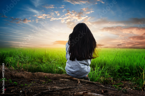 A little girl sits in the middle of a field watching the sun go down all alone.